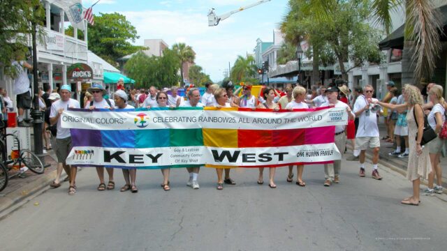 A diverse group of people walk down Duval Street in Key West holding a large rainbow-colored banner that reads “Original Colors – Celebrating Rainbow25 All Around the World – Key West – One Human Family.” Behind them, people begin to unfurl the mile-long Sea to Sea Rainbow Flag as spectators line the sidewalks.
