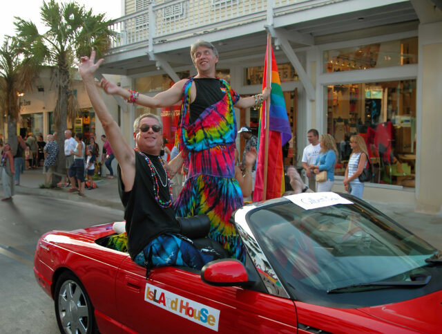 Gilbert Baker and Cleve Jones ride together in a bright red convertible labeled “Island House” during the 2003 Key West Pride Parade. Gilbert wears a rainbow toga and waves joyfully with a Pride Flag in hand, while Cleve, in sunglasses and rainbow beads, celebrates beside him. Spectators line Duval Street behind them.