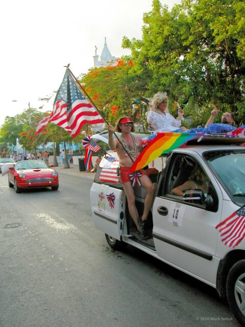 A shirtless man in red shorts with star-spangled bows, boots, and beads leans from the open door of a decorated white van in the Key West Pride Parade on June 14, 2003. He waves a large American flag while a rainbow flag hangs across the side of the van. Inside, a drag performer in a blond wig and white dress smiles and gestures toward the cheering crowd. Another parade car follows behind on Duval Street, framed by tropical trees and the spire of St. Paul’s Episcopal Church.